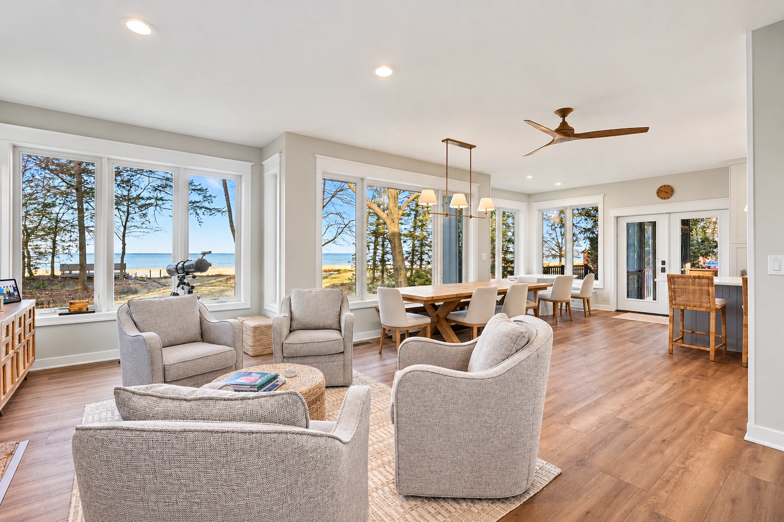 Bright all-season sunroom with gray armchairs, a long dining table, and wide windows overlooking a beach.