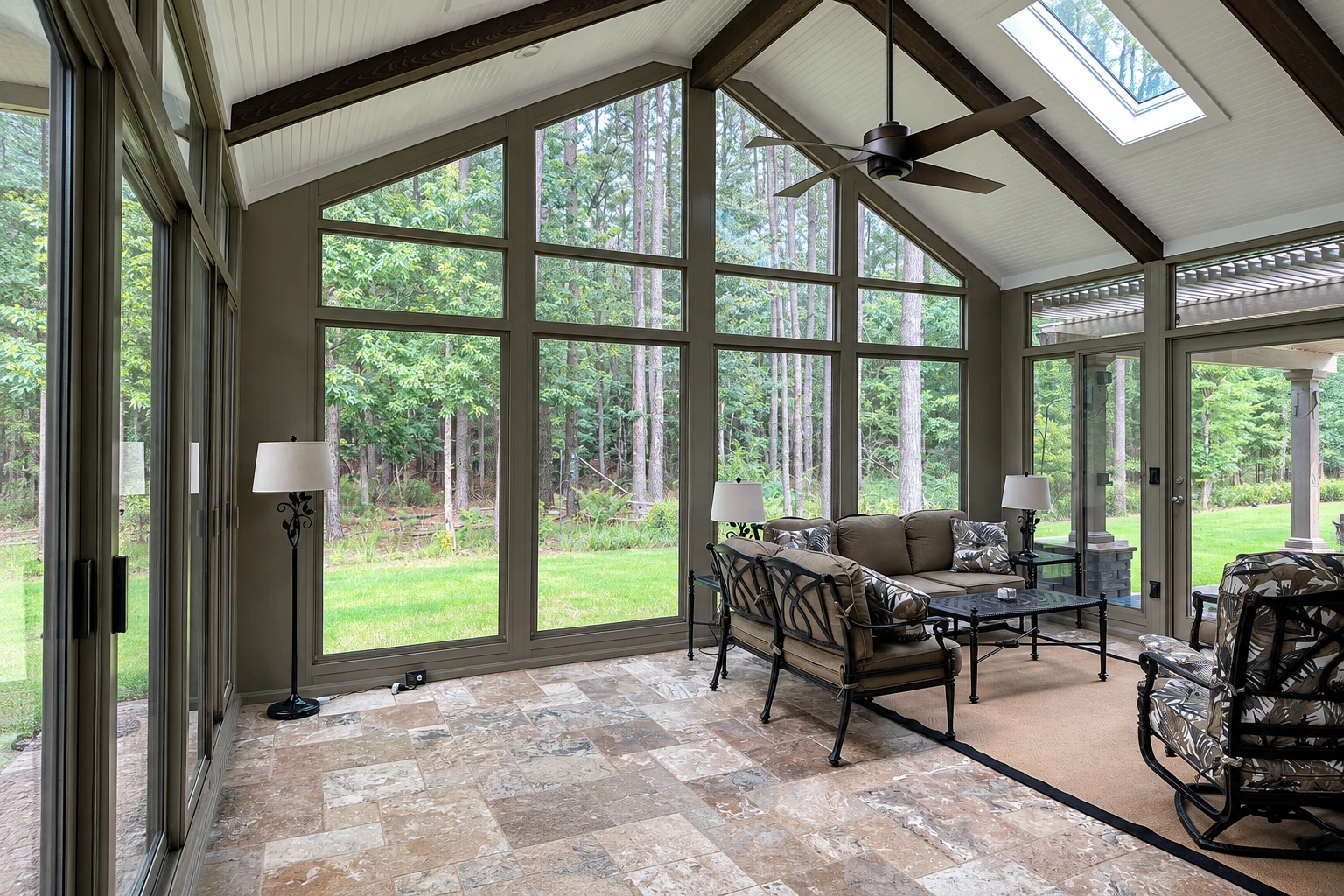Four-season sunroom with vaulted white ceiling, dark exposed beams, expansive windows, and a seating area facing woods.