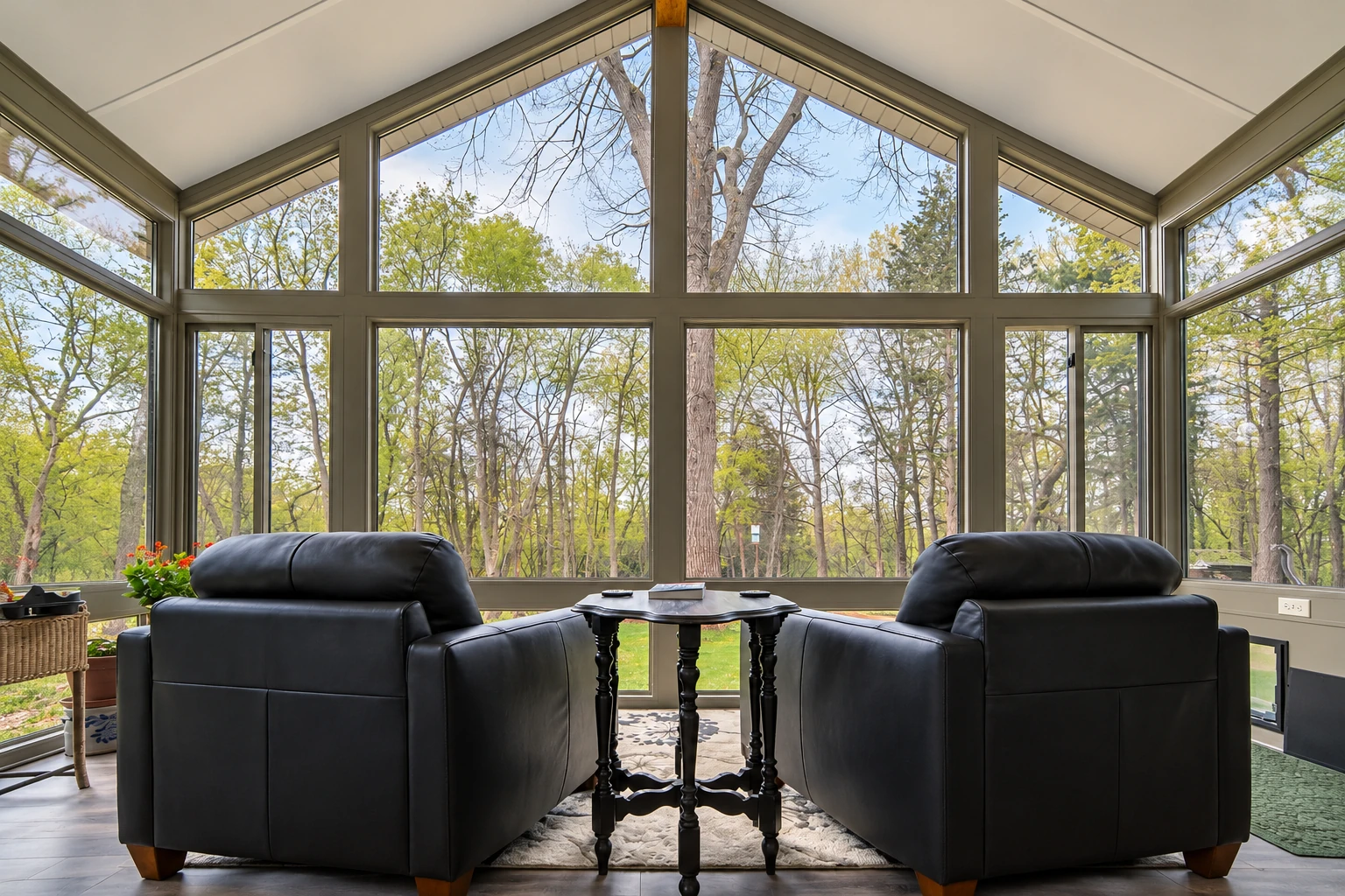 Custom sunroom with a peaked glass wall, two black leather chairs, and a small table facing a wooded view.