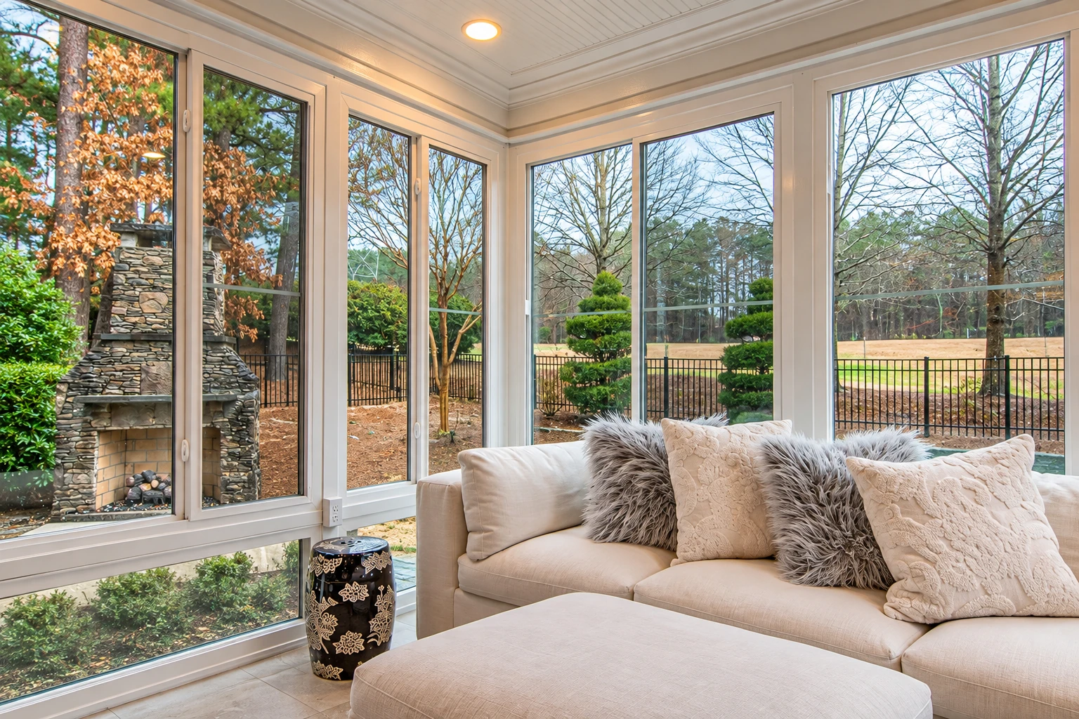 Four-season sunroom with tall white-framed windows, a cream sofa, and views of bare trees and a stone fireplace outside.