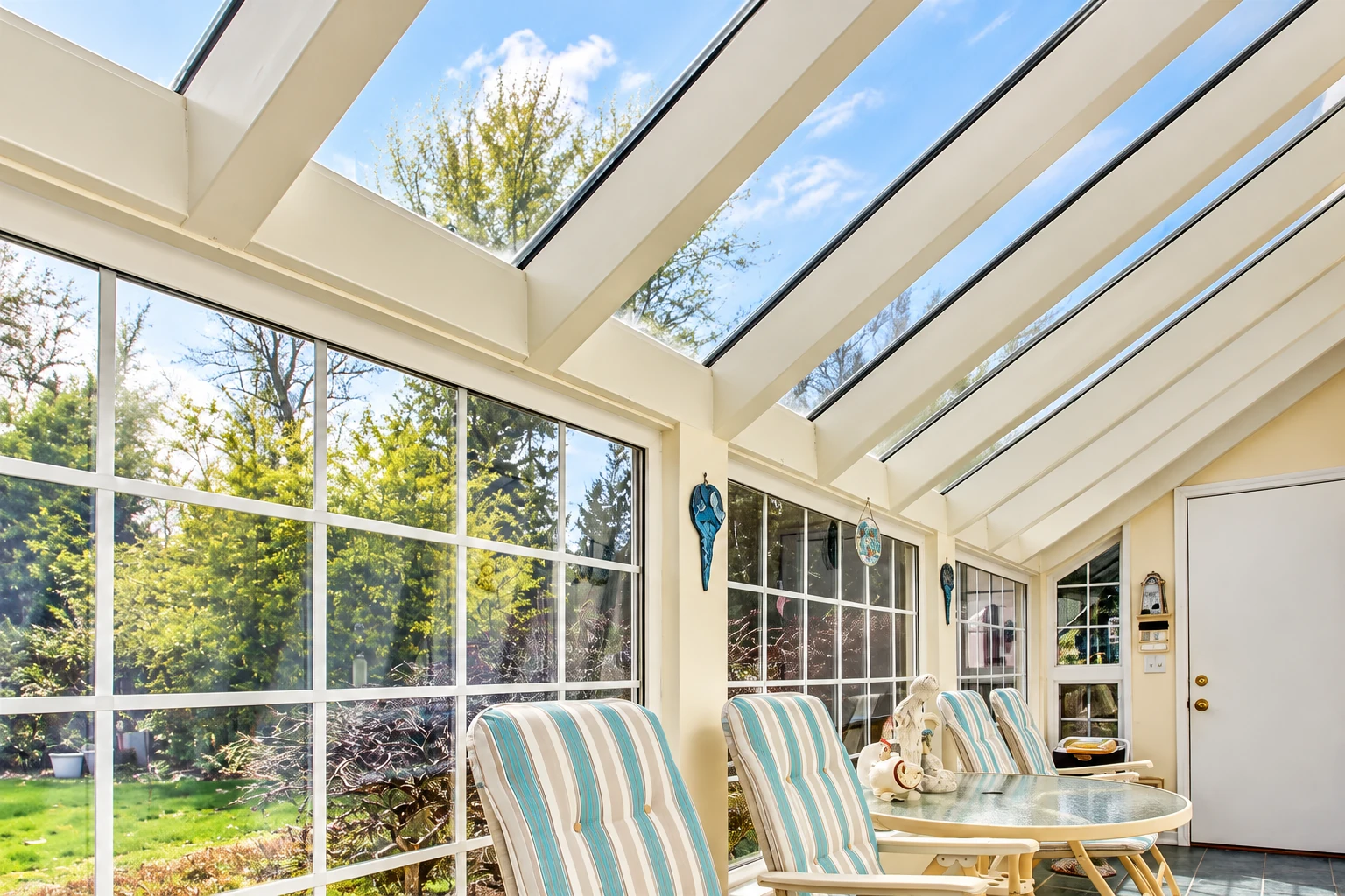 Solarium with a sloped glass roof, tall windows, and striped lounge chairs beside a round table.