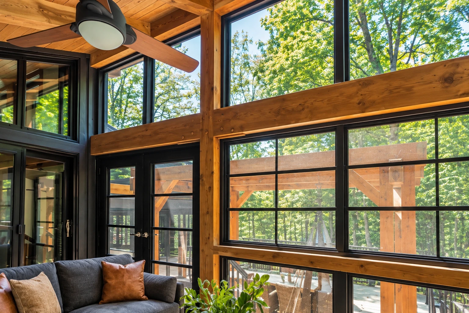 Sunroom addition with warm timber beams, black-framed windows, a ceiling fan, and a gray sofa overlooking leafy trees.