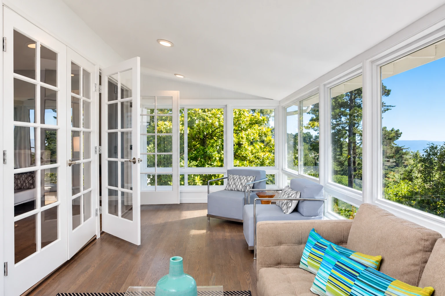 Bright sunroom with white French doors, large windows, and blue and beige seating overlooking leafy trees.